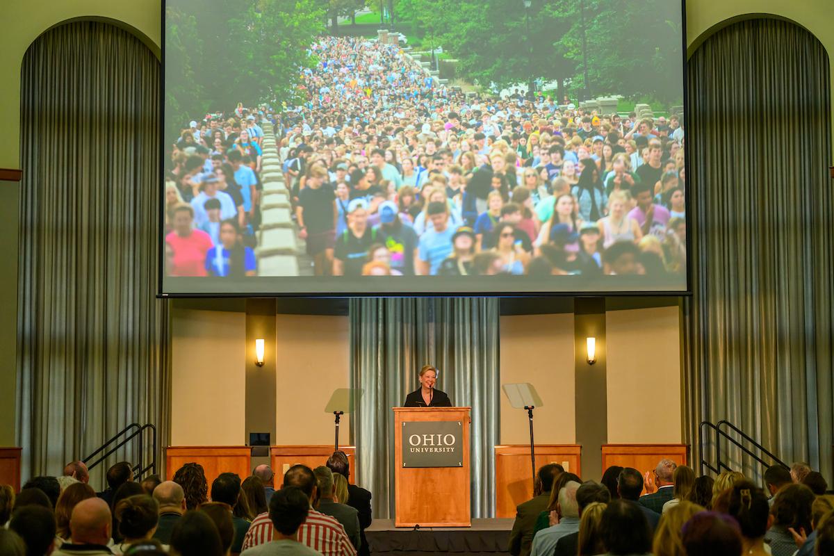 President Lori Stewart Gonzalez speaks at the podium in Walter Hall during the State Of the University Address. Above her on the video screen is an image of OHIO students walking up Richland Avenue during a Welcome Week event.