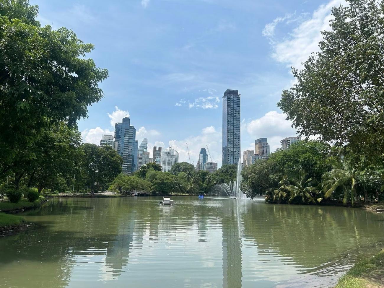 An image from Thailand of a lake with trees surrounding it and buildings in the background, the sky is blue overhead