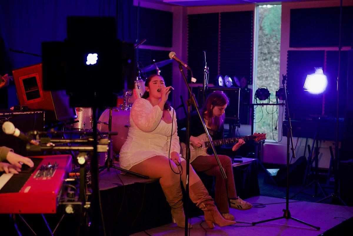 An individual sings into a microphone while sitting on a stage and people play musical instruments nearby