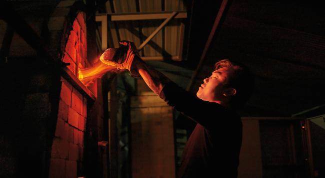 flames shoot from a small gap as a graduate student wearing gloves removes a brick from a kiln to check on its contents