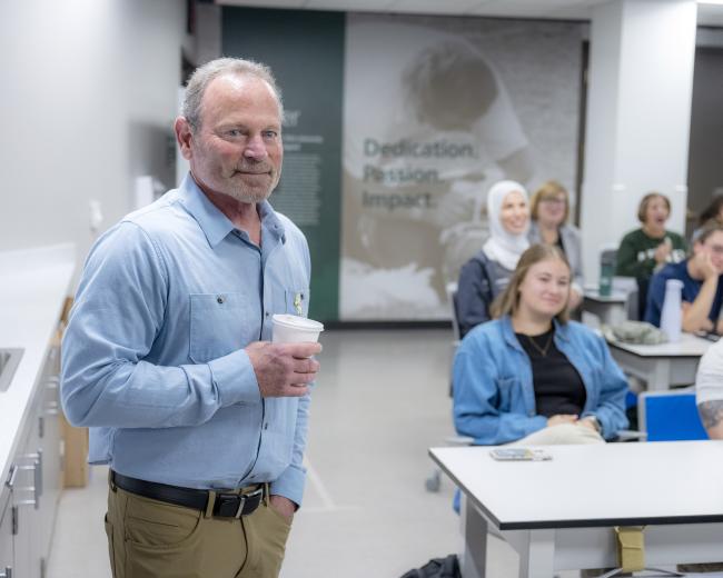 a man with a short gray beard and hair in a blue button-down shirt and khakis holds a coffee cup in front of a group of students sitting at desks