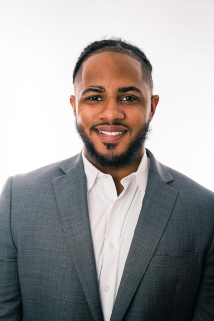 a Hispanic man with short black hair and a beard and mustache wearing a gray suit and white oxford shirt