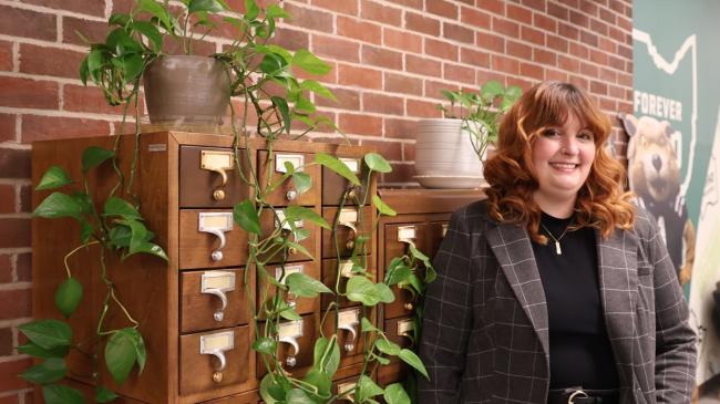 Haley Shaw stands next to a card catalog that hs plants on it, and there is a Forever OHIO banner on a wall in the background