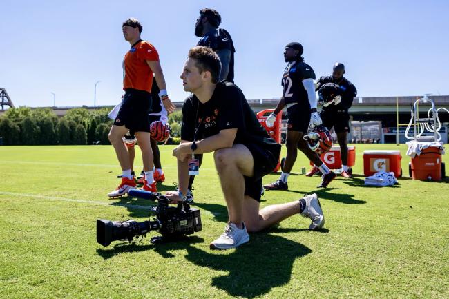 Jack Cowman kneels beside Cincinnati Bengals quarterback Joe Burrow on the field, camera in hand