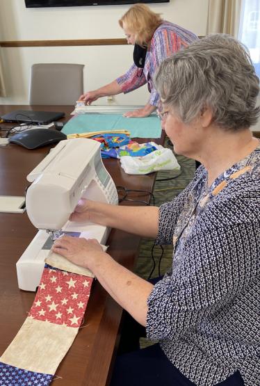 One lady works on sewing together quilt pieces while another in the background works on cutting fabric