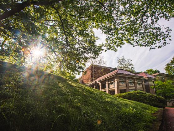 The sun rises behind a lush green hill and tree, with the Scripps Hall in the background