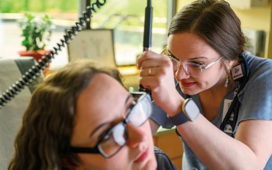an instructor wearing glasses and scrubs demonstrates the use of an otoscope on a student