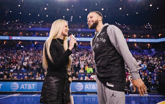 sports reporter Allie LaForce holds a mic and speaks to basketball player Stephen Curry in a packed basketball arena