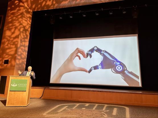 Dr. Ariel Armony gives his presentation in the Baker Theater, while a slide on the wall behind him shows a human hand touching a robotic hand