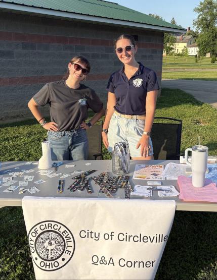 Gracie Vaughn and Hannah Wynne stand behind a table at an outdoors tabling event in Circleville