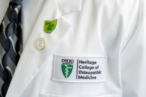 The Heritage College of Osteopathic Medicine logo is shown on a white coat, worn by a doctor. The coat also has a pin that says Care Leads Here