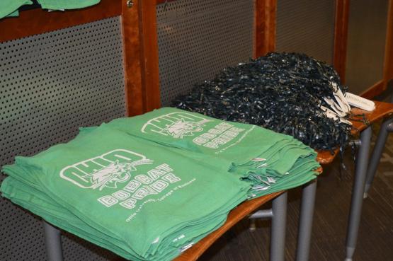A stack of OHIO shirts that say "Bobcat Pride" and have an OHIO logo on them and a stack of pom poms
