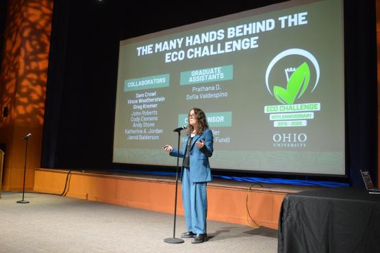 An individual gives a presentation on the stage at the Eco Challenge, with an Eco Challenge graphic on the screen behind her. The graphic says "The Many Hands Behind The Eco Challenge."