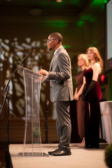 A speaker talks at the podium while two students standing on stage watch during the Leadership Awards Ceremony in the Baker University Center Ballroom.