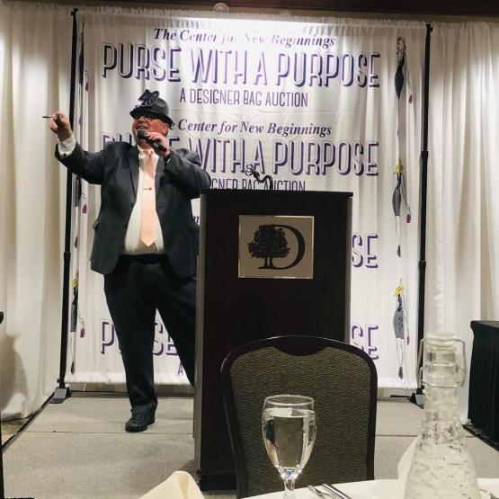 Nate Strum is shown wearing a suit and hat while holding a microphone and speaking near a lecturn at an event. I banner behind him says "The Center for New Beginnings,  Purse With A Purpose, A Designer Bag Auction"