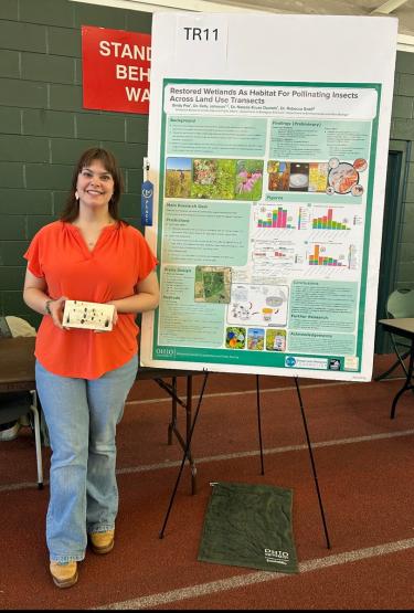 Emily Fox stands next to her research poster and also holds a small box at the 2026 Student Expo