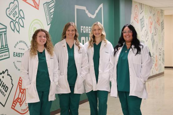 4 Nursing Students dressed in green scrubs, wearing white coats standing beside one another