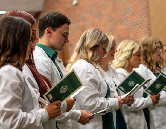 Row of students in white coats looking at program during pinning ceremony