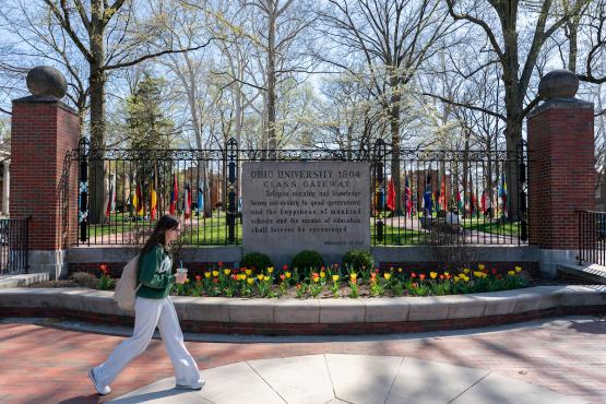 An OHIO student walks in front of the College  Gate, which has colorful flowers in front of it and colorful trees behind it, on a spring day