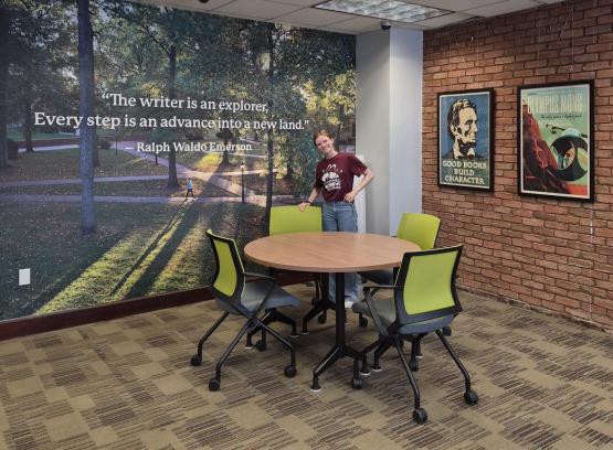 An OHIO student stands by a round table at the renovated Writing Commons. The table is in front  of a wall that has an image of the College Green and the words "The writer is an explorer. Every step is an advance into a new land - Ralph Waldo Emerson"