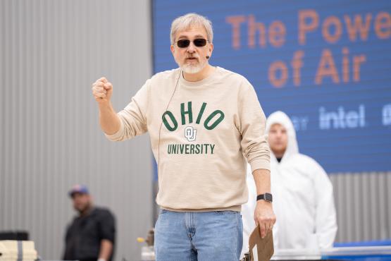 A speaker wearing an Ohio University shirt talks to the crowd at the Science Day event while an OHIO representative in a lab coat and hood is in the background.