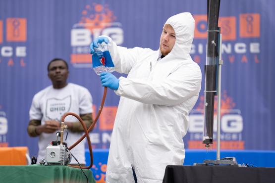 An OHIO student in a lab coat and hood holds a large soda bottle up to a hose as part of an experiment, while an OHIO representative wearing an Ohio University shirt watches in the background at the Science Day event