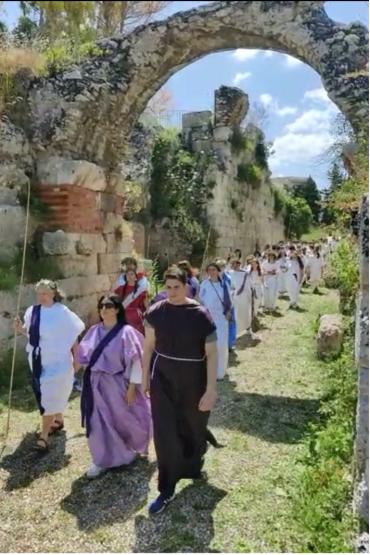 A group of people in traditional Greek dress walk in a line beneath a stone archway in the open air