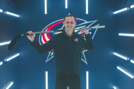 Nathan Wiseman stands in front of a Columbus Blue Jackets backdrop with a hockey stick slung over his shoulders