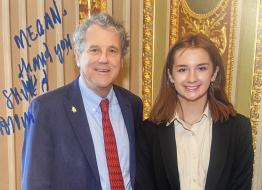 Senator Sherrod Brown poses for a portrait with a college student, both are wearing blazers and smiling