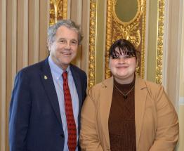 Senator Sherrod Brown poses for a portrait with a college student; both are wearing blazers and smiling
