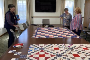 Three women stand around a board table, with two unfinished quilts resting on the table