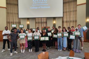 A group of students stand in a large reception room posing for a picture, holding certificates