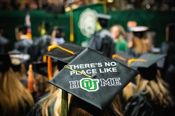 An OHIO student is shown wearing a graduation cap that says "There's No Place Like HOUme at Fall Commencement 2023