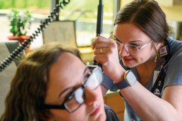an instructor wearing glasses and scrubs demonstrates the use of an otoscope on a student