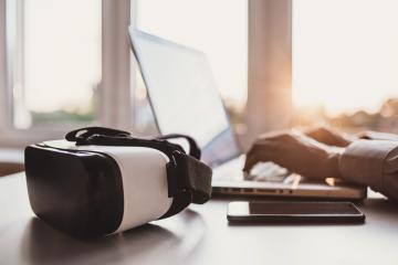 Virtual reality headset on a desk with someone on a laptop behind it.