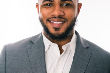 a Hispanic man with short black hair and a beard and mustache wearing a gray suit and white oxford shirt