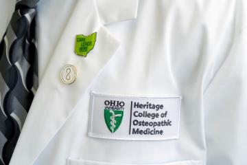 The Heritage College of Osteopathic Medicine logo is shown on a white coat, worn by a doctor. The coat also has a pin that says Care Leads Here
