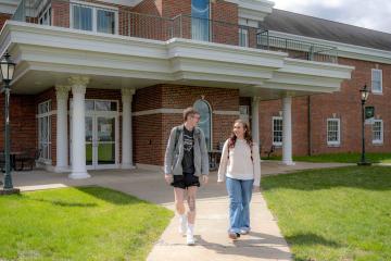 A male and female walking next to each other walking down a sidewalk, outside the rear entrance to Ohio University Eastern Shannon Hall