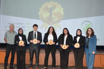 The winners of the Eco Challenge, along with advisors, hold trophies while standing on stage at the Eco Challenge, and a 1st Place graphic is shown behind them