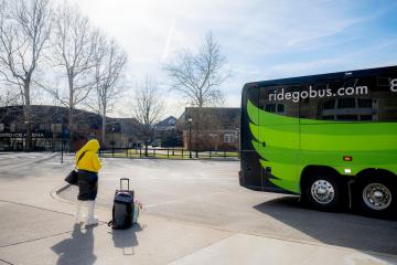An OHIO student with a suitcase  waits outside of a GoBus parked near Baker University Center, Bird Arena and the Aquatic Center