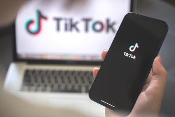 a person holding a cell phone with the TikTok logo, in the background a laptop with the same logo