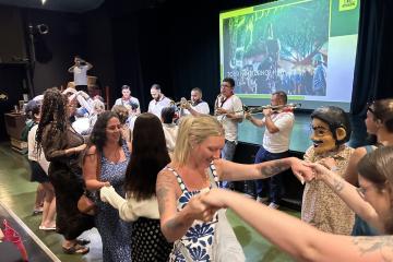 Individuals dance together while music  plays and one person wears a costume