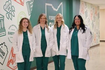4 Nursing Students dressed in green scrubs, wearing white coats standing beside one another