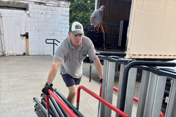 One member of the Moving and Surplus Team moves a cart filled with poles and other items outside while another member of the team moves items in the back of a moving truck