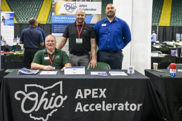 Three individuals sit and stand behind a table that has an Ohio APEX Accelerator banner on it at the Ohio Business Matchmaker event