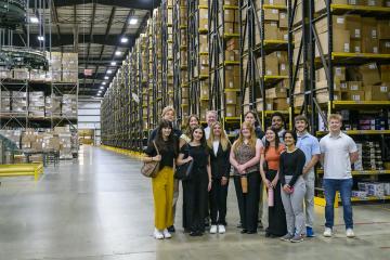 A group of student interns stand together inside a very large warehouse filled with shelves that are full of boxes