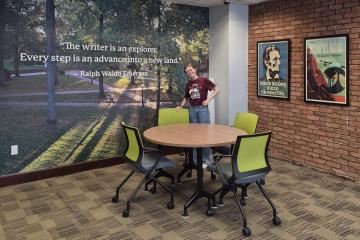 An OHIO student stands by a round table at the renovated Writing Commons. The table is in front  of a wall that has an image of the College Green and the words "The writer is an explorer. Every step is an advance into a new land - Ralph Waldo Emerson"