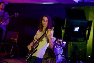An individual plays guitar while another person plays a guitar in the background