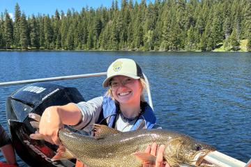 A woman wearing a baseball hat and standing in a boat with pine trees in the background holds a freshly-caught fish out for the camera