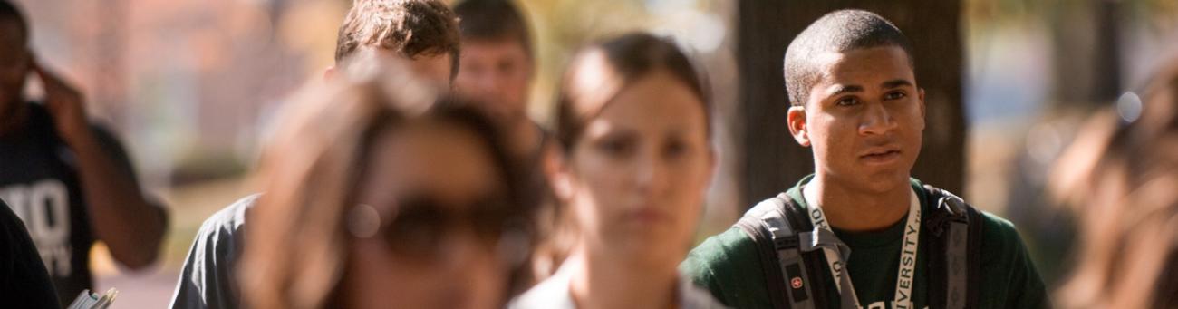College student with backpack walking on busy campus pathway on sunny fall day, with other students visible in background